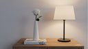 A single white preserved rose in a slim white bud vase beside a table lamp on a wooden desk