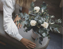 A florist arranging preserved white roses and eucalyptus in a ribbed white vase