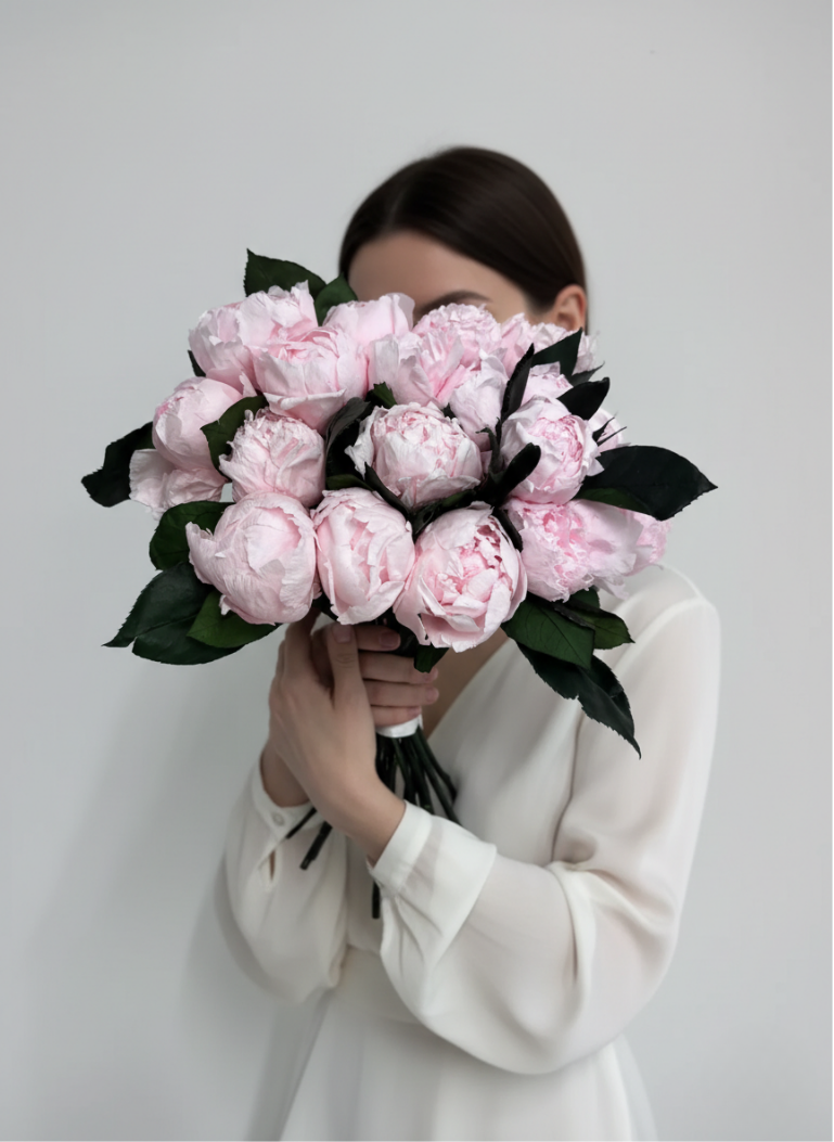 A woman holds a large bouquet of nordblooms preserved pink peonies and dark green leaves, covering her face. She wears a white blouse and is against a light grey background. The flowers are in focus, filling much of the frame.