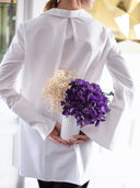 A woman holding a vase of purple preserved hydrangea and dried flowers behind her back