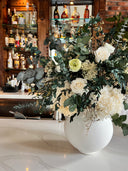 A large preserved arrangement of white blooms and eucalyptus in a white globe vase on a bar counter